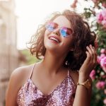 Outdoor close up portrait of young beautiful happy smiling curly girl wearing stylish heart gradient sunglasses, pink sequin blouse, watch. Model posing near blooming roses. Summer fashion concept