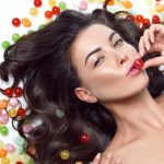 Beautiful young sexy brunette woman dark long hair evening makeup perfect shape tanned body accessories jewelry eating sweet candy candies different colors and flavors organic and natural diet fitness